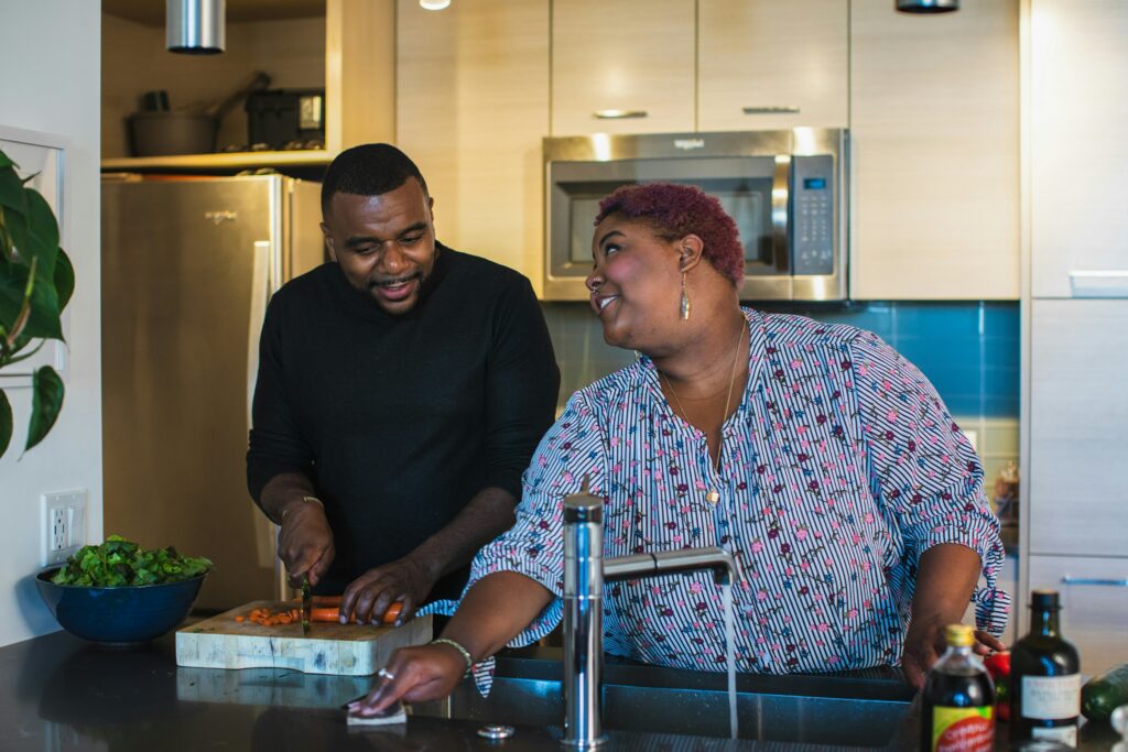 This charming image captures a couple in their kitchen, immersed in the joy of cooking together. Laughter lights up their faces as they collaborate on a shared culinary adventure. The warmth of their happiness is palpable, turning the kitchen into a lively space filled with love, shared moments, and the aroma of delicious food as they are elevating relationships through self-care.