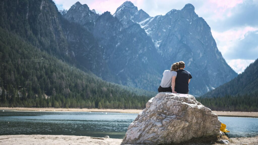 Against the backdrop of a serene lake, a couple sits embraced on a rock, captivated by the majestic mountains in the distance. Their connection is evident in the tender embrace, and as they gaze across the water, a shared sense of tranquility and awe envelops the scene as they are elevating relationships through self-care.